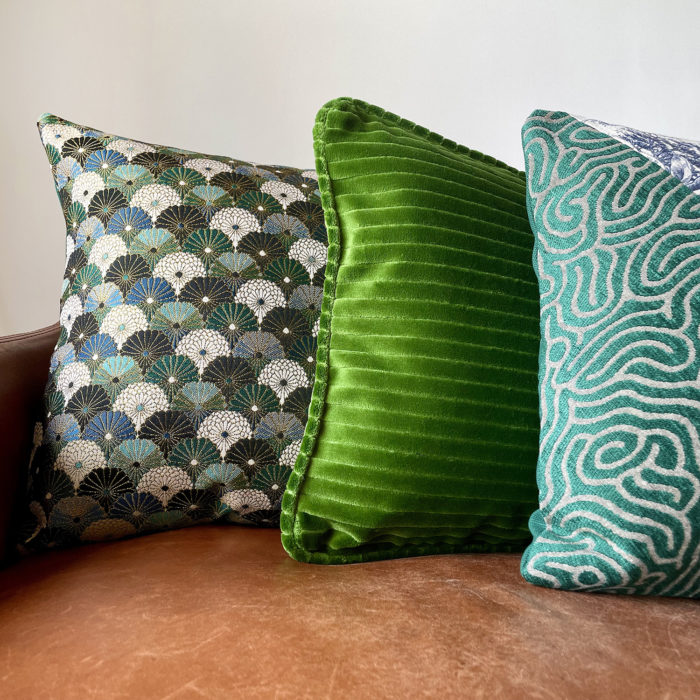 Coussins décoratifs verts sur canapé en cuir, mêlant velours côtelé vert intense, motif éventails art déco et tissu graphique turquoise.